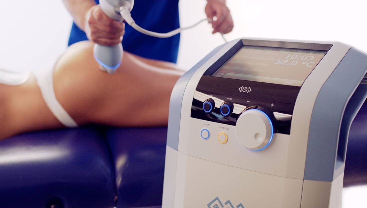 An Emsculpt NEO machine is in the foreground and a body contouring procedure is taking place on a woman's buttocks in the background